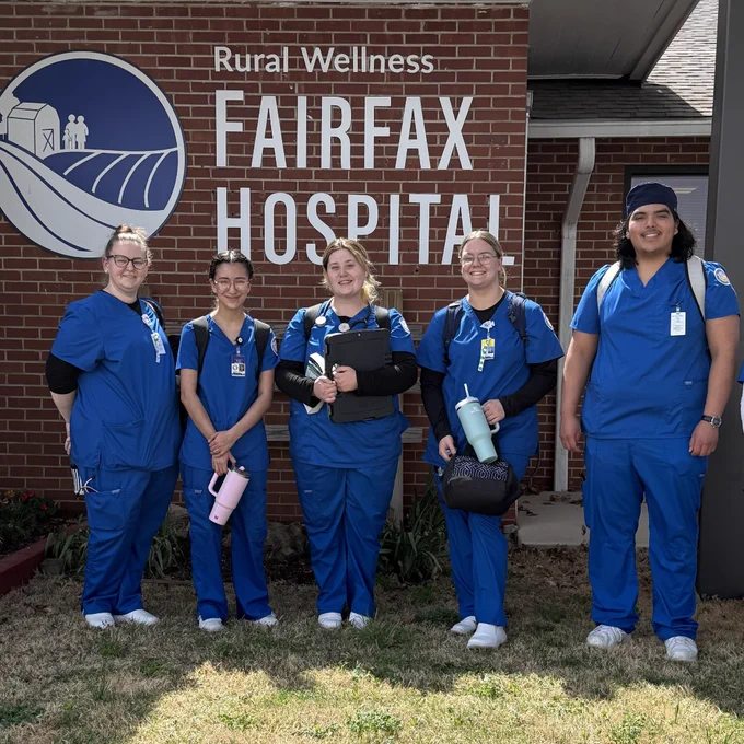 Group of six healthcare workers in blue scrubs standing outside Fairfax Hospital under a Rural Wellness sign.