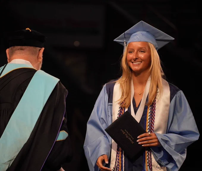 A female graduate with long blonde hair smiles onstage after receiving her diploma.