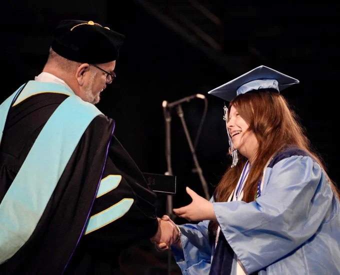 A side view of a female graduate with long brown hair sharing her principal’s hand and receiving her diploma onstage.