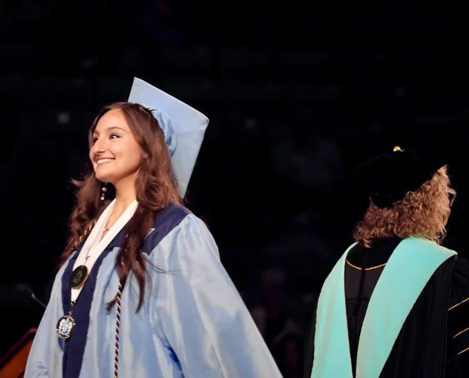 A side view of a female graduate with wavy brown hair smiling brightly while walking onstage at commencement.