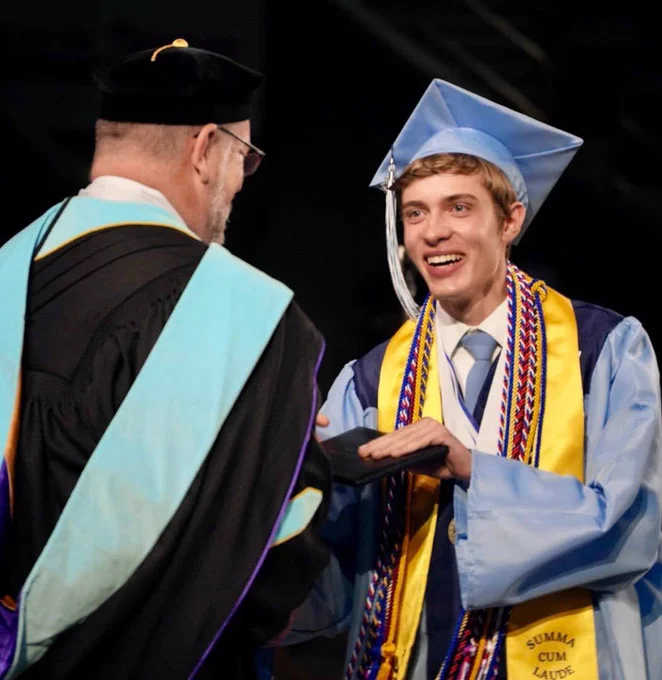 A male graduate smiles widely and brightly as he receives his diploma from his high school principal.
