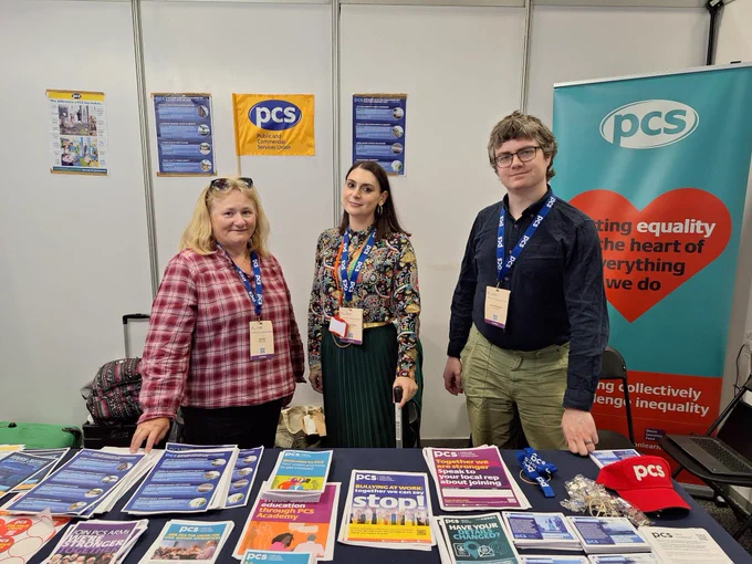 The photo shows PCS staff Gail, Maria and Liam stood in front of a table holding various PCS leaflets, lanyard and hats with a backdrop of PCS posters and a pull-up stand which reads PCS (partially obscured) putting equality at the heart of everything we do. 
