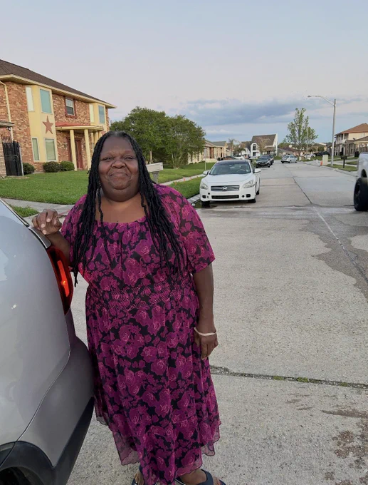 a photo of maw maw linda, a dark skin black woman with black locs sitting on her shoulders in a fuchsia and black floral printed sundress that rests at her ankles. she is smiling and leaning against a gray suv. in the background there is a neighborhood of homes.