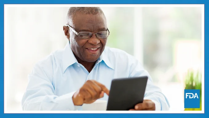 A person smiling while using a tablet, seated at a table with a cup and a small plant. The FDA logo is visible in the bottom right corner.