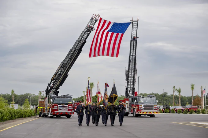 Firefighters march under a large American flag held aloft by two firetruck ladders. The scene is formal and patriotic.