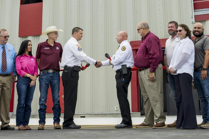 A group of people, including fire department officials in uniform, gather for a formal ceremony. The central focus is on two officials shaking hands, signifying a formal agreement or celebration. The mood is professional and congenial.