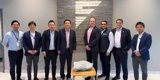 Eight men in business attire standing in a row inside a modern office lobby with a large geometric logo on the wall behind them.