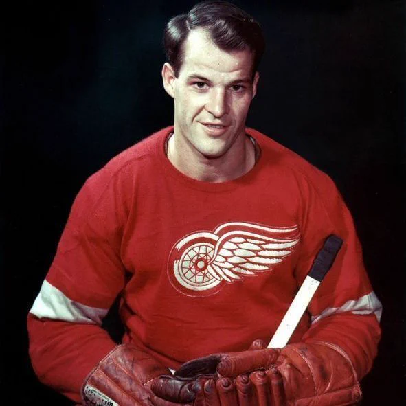 Gordie Howe poses in a red Detroit Red Wings jersey with the winged wheel logo on the chest. He is wearing red hockey gloves and holding a hockey stick in front of a dark background.

