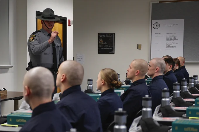 A uniformed Pennsylvania State Police Trooper speaks in front of a classroom with seated cadets