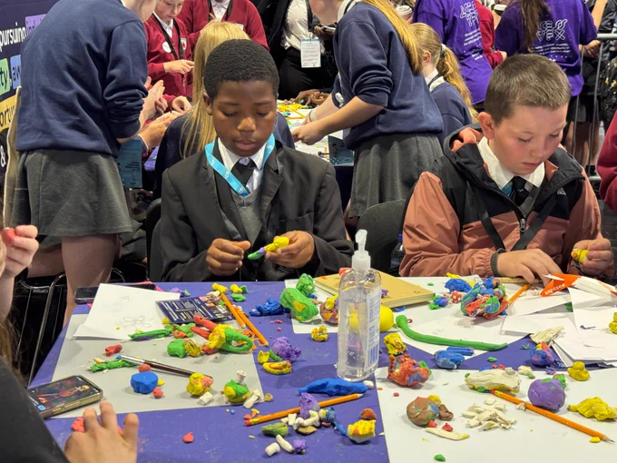 A group of young students at a table are engaged in crafting activities using colorful modeling clay. The table is cluttered with clay pieces and tools. In the background, more students participate in similar activities in a busy environment.