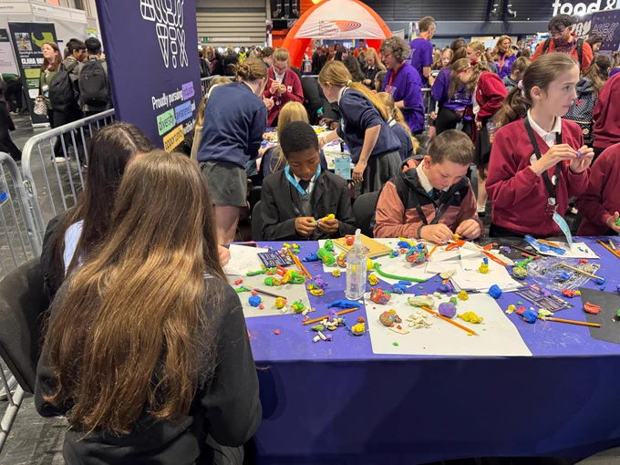 A group of people are engaged in a creative activity at a table filled with colourful art supplies, including clay figures and crafting materials.