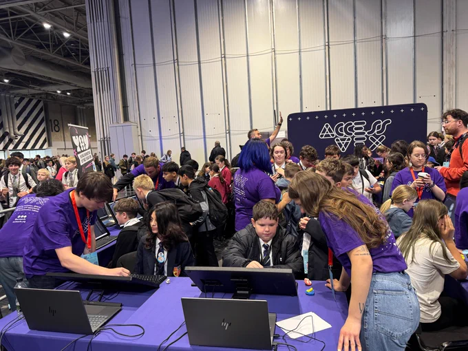 A crowded event gathering at an Access VFX-branded booth. Many attendees are engaged with various computer screens, assisted by individuals wearing purple shirts. The background features a large Access VFX sign.
