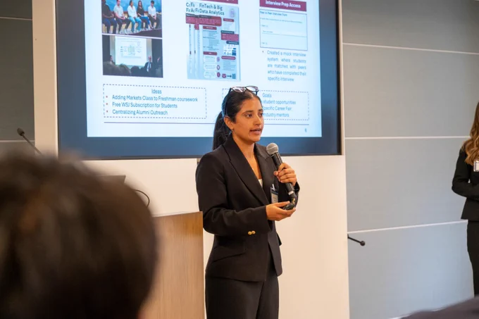 A Pamplin student presents to an audience during the Hokies on Wall Street program, sharing ideas and insights while standing in front of a presentation screen in a professional meeting space.