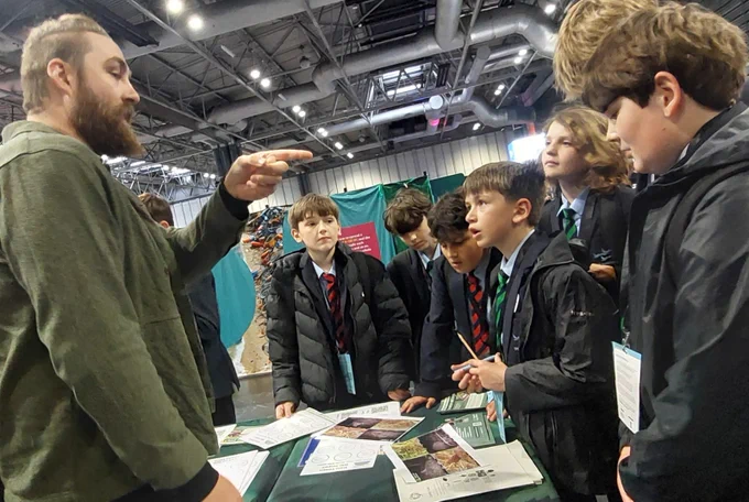 A group of students in school uniforms attentively listen to a person speaking at a table filled with brochures and papers. 