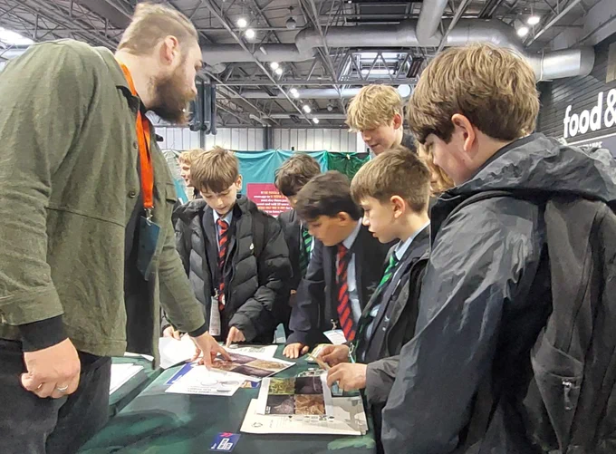 A group of people are gathered around a table with brochures at The BIg Bang Fair. One individual is explaining something to the group.
