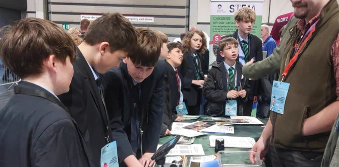 A group of young individuals in school uniforms gather around a table at an event. They appear engaged, looking at various materials spread out before them. Posters in the background mention "STEM" and "Careers on Our Property."