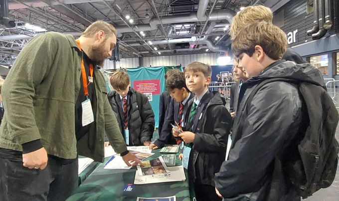 A group of young students in uniforms is gathered around an exhibitor at a booth during an event at the NEC Birmingham. The exhibitor is discussing materials displayed on the table. The event hall is spacious, with visible signage in the background.