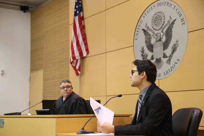 Student participating in a mock trial competition speaks into a microphone from the witness stand inside a courtroom. Photo Credit: Arizona Bar Foundation. 