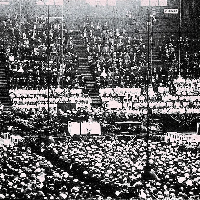 Black-and-white historic photograph of a packed indoor arena or hall filled with a large crowd. Rows of spectators sit on steep wooden bleachers, while a dense audience fills the floor in front of a decorated stage or platform. Several people sit at long tables near the front, with bunting, plants, hanging lights, and draped fabric around the stage area. Stairways, railings, an Exit sign, and a No Smoking sign are visible in the background.

