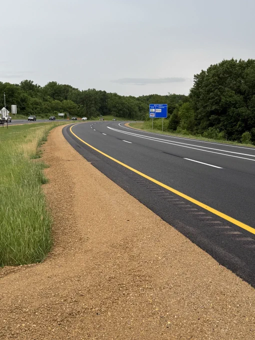Curved highway with fresh asphalt and clear lane markings surrounded by trees under a cloudy sky.