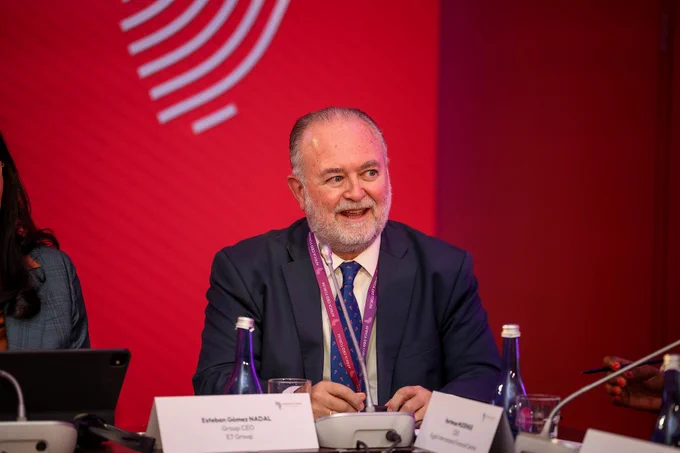 A business leader sits at a conference panel with a microphone and a nameplate indicating a discussion setting. The atmosphere is professional, set against a red backdrop.
