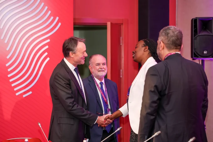 A group of business professionals engage in conversation and handshake, indicating a formal, positive interaction during a conference. The setting suggests networking and collaboration.
