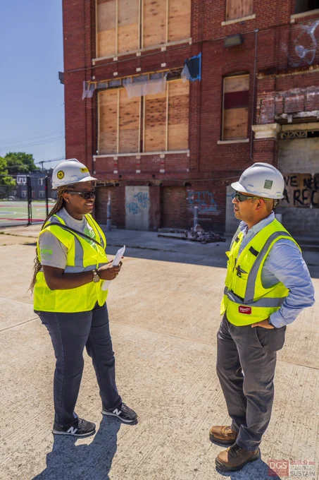 Two employees wearing hard hats and neon yellow safety vests discuss plans at an outdoor site on a sunny day.