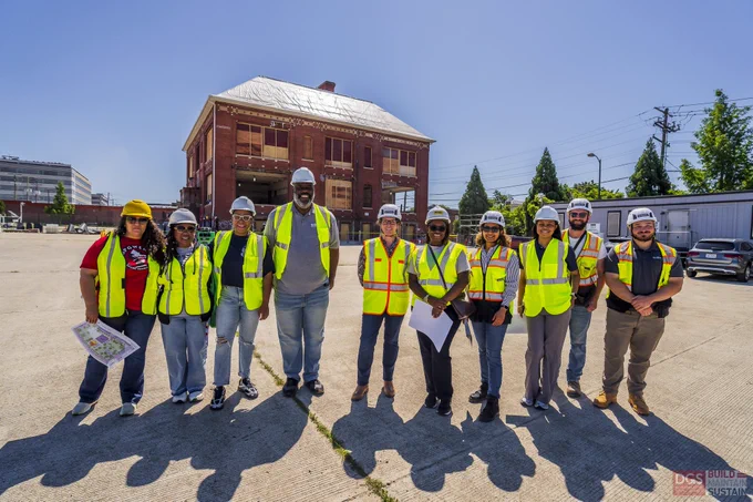 Group of employees and community members wearing safety vests and helmets standing together on a sunny site in front of a brick building.