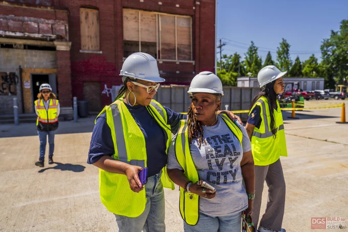 Two people, DGS employee and an ANC Comissioner in yellow safety vests and white helmets stand together outside an industrial building on a sunny day.