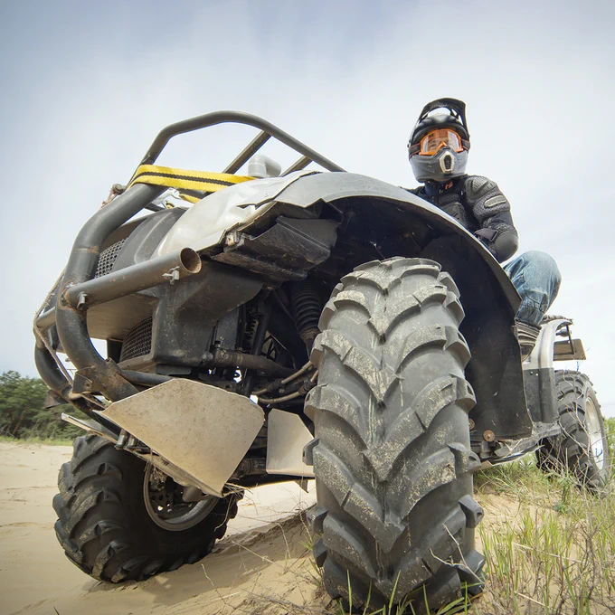 A person wearing a helmet is sitting on a large off-road ATV in a sandy outdoor setting. The vehicle's rugged tires are prominently visible.