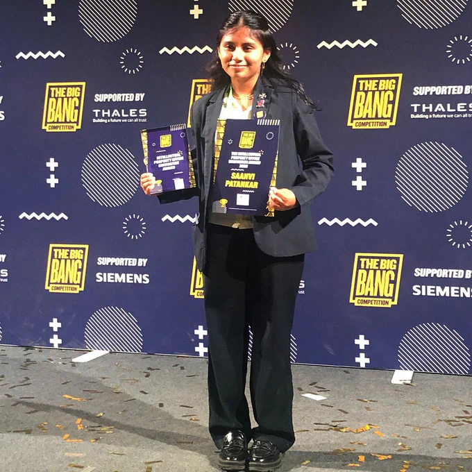 Person holding two awards on stage at The Big Bang Competition event with sponsor logos in the background.