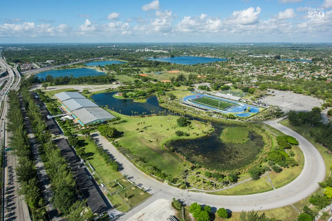 Aerial view of a sprawling park and complex. On the left, a multi-lane highway runs beside large buildings. The park features multiple blue lakes, green fields, a stadium with a blue track and 'HOME OF THE WILDCATS' football field. Residential areas are visible in the distance. A logo in the top right reads: MIAMI-DADE 250 America.
