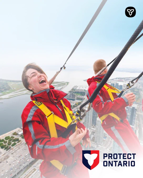 Three people in safety harnesses stand on the edge of a tall building, overlooking a city and waterfront far below.