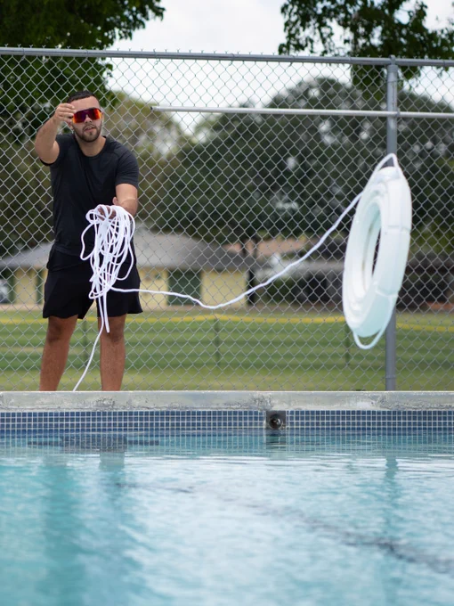 A person next to a swimming pool is throwing a ring buoy attached to a rope toward the water. There is a chain-link fence and trees in the background.