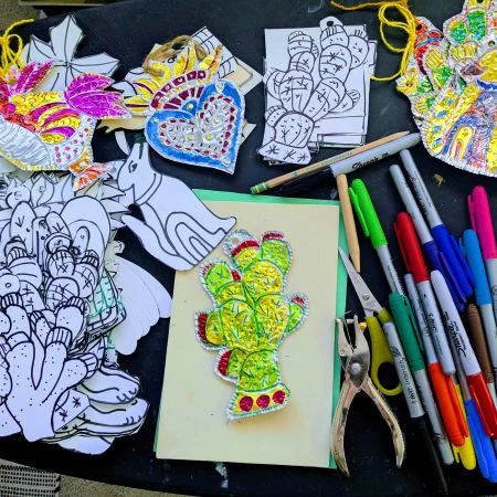 Colorful cactus-shaped paper craft decorated with markers surrounded by black and white cutouts and art supplies on a table.