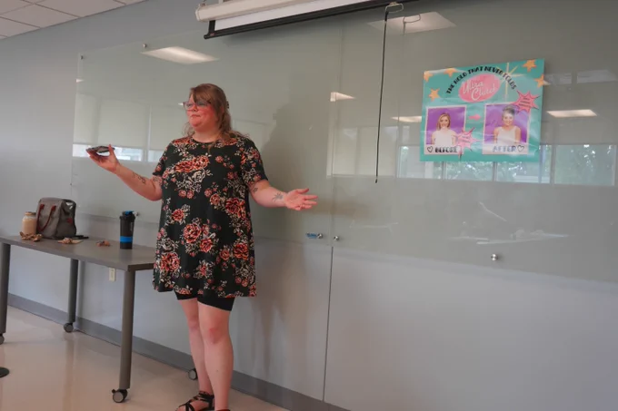 Woman in a floral dress giving a presentation in a classroom with a colorful poster on the wall behind her.