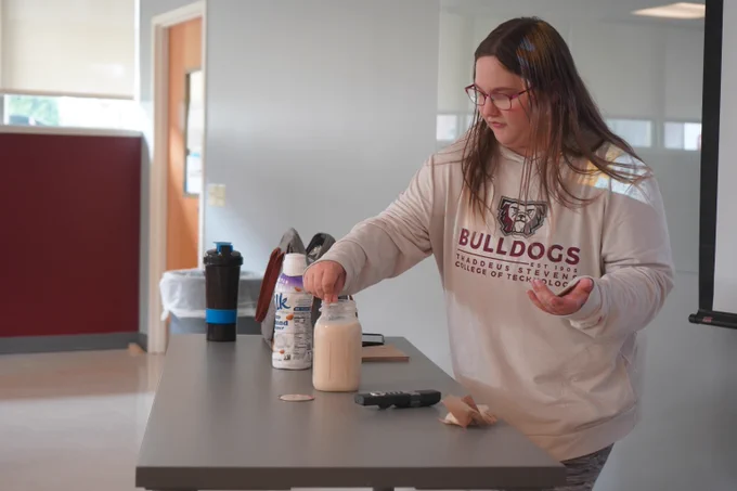 Woman in Bulldogs sweatshirt stirring a drink in a jar on a table with various bottles nearby.
