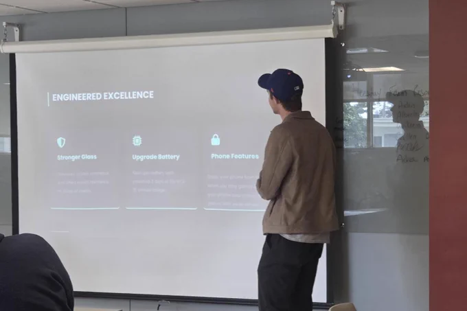 Man presenting a slide titled 'Engineered Excellence' highlighting stronger glass, upgraded battery, and phone features.