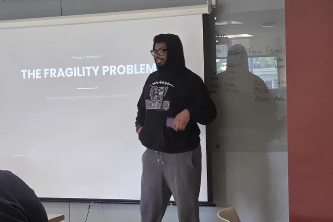 Person wearing a black hoodie and gray pants stands in front of a projection screen displaying a presentation titled 'THE FRAGILITY PROBLEM'.