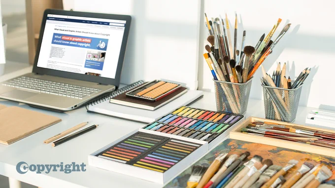 Table full of art supplies - pencils, paintbrushes, paints, pastels, notepads. Laptop on the left open to a web page. 