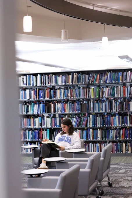 Person reading a book while seated in a modern library with tall shelves filled with books behind them.