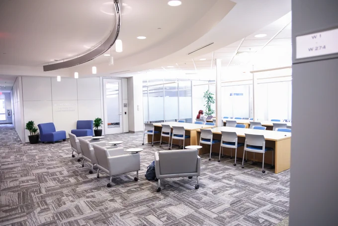 Modern office lounge area with gray armchairs and blue chairs around a long wooden table under bright ceiling lights.