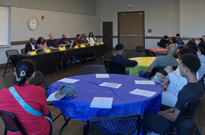 Panel of seven speakers seated at a long table addressing an audience in a conference room with round tables covered in colorful cloths.