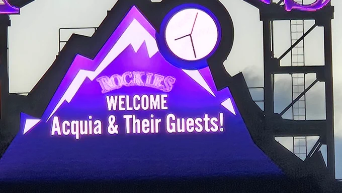 Illuminated scoreboard at a Rockies stadium welcoming Acquia and their guests with a mountain and clock design.