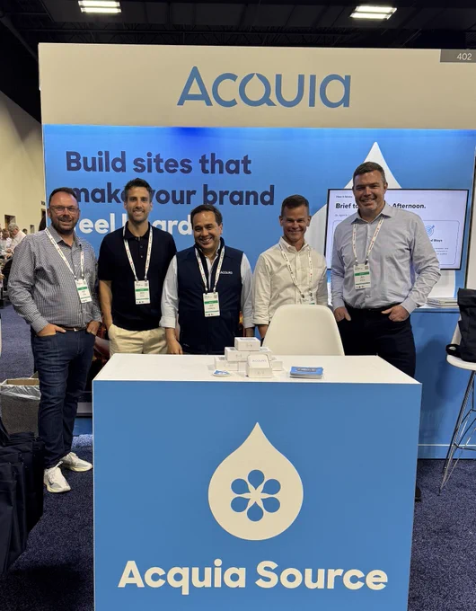 Five men standing behind a blue and white Acquia Source booth at a conference, promoting website building services.