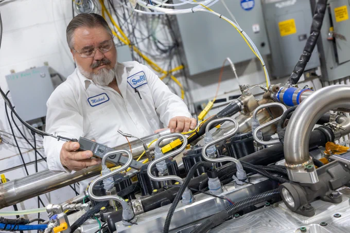 SwRI staff member in a white lab coat using diagnostic equipment on a complex engine system inside an industrial workshop.