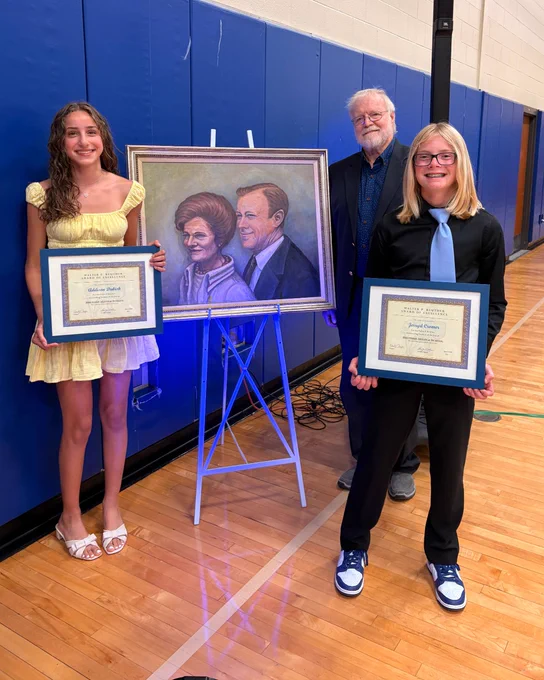Two students holding framed certificates stand in a gymnasium next to a portrait of Walter Reuther.