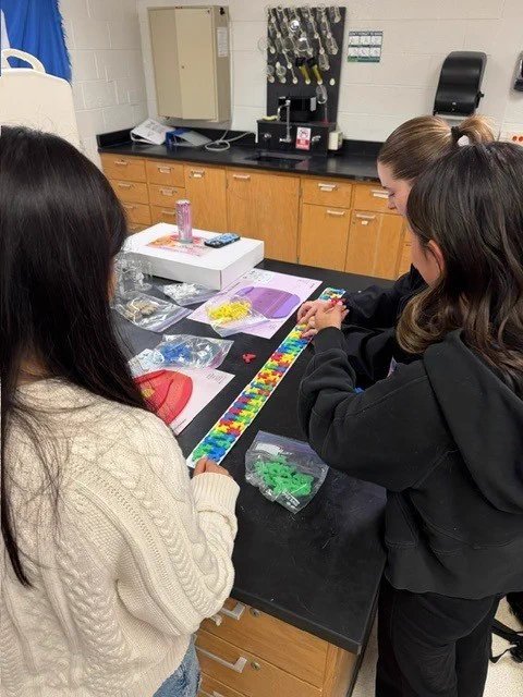 Two students working on a molecular modeling project at a classroom lab table with supplies spread out.