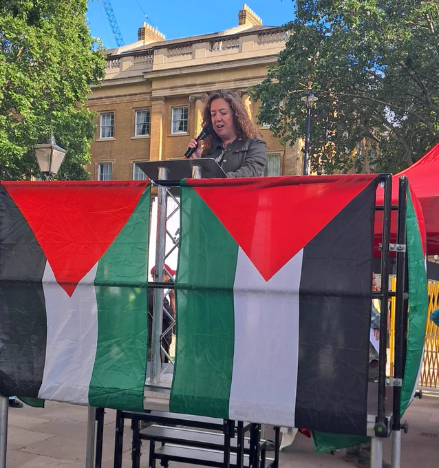 The photo shows PCS General Secretary Fran Heathcote holding a microphone standing at a lectern on a stage draped with Palestine flags which feature three equal horizontal bands of black, white, and green, overlaid with a red triangle based at the hoist