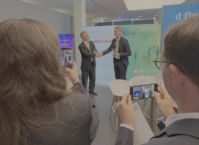 Dr. Frank Schlottmann (msg) and Niels Lindenthal (OpenProject) marking the announcement of the partnership between msg and OpenProject with a handshake at the Zukunftskongress Staat & Verwaltung 2026 in Berlin, while attendees capture the moment on their smartphones.
